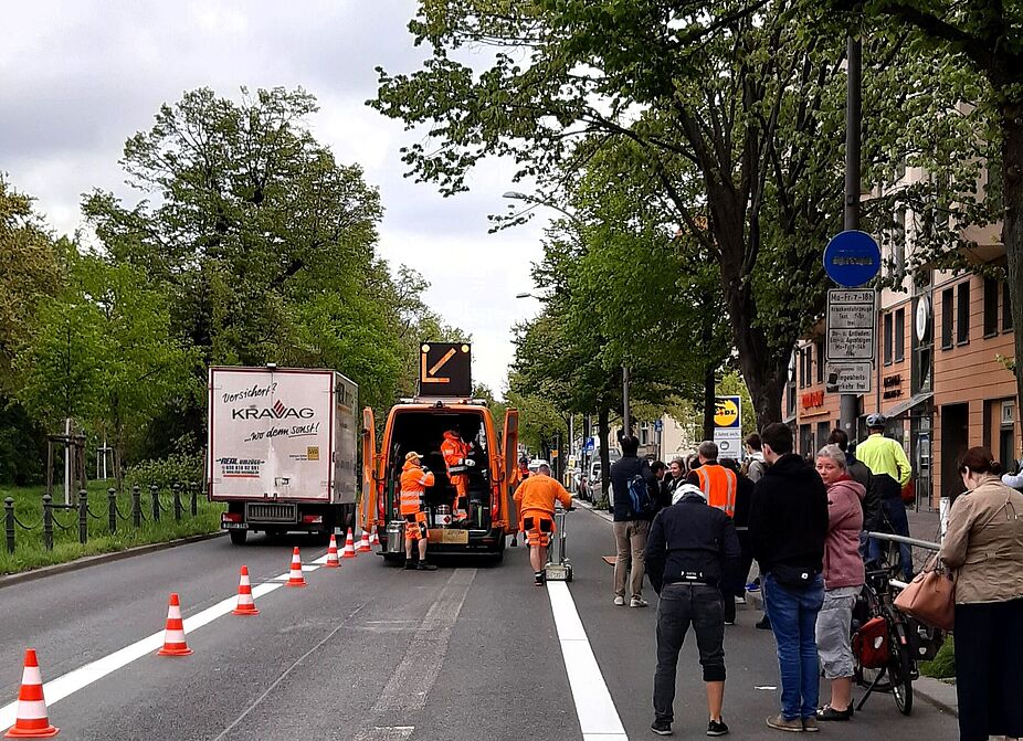 April 2024: Markierungsarbeiten an der Schöneberger Hauptstraße Mehrere Menschen schauen sich auf der Hauptstraße zwischen Eisenacherstraße und Martin-Luther-Straße Markierungsarbeiten an. Zu sehen sind auch ein Baufahrzeug und ein frisch gemalter weißer Streifen auf der Fahrbahn.