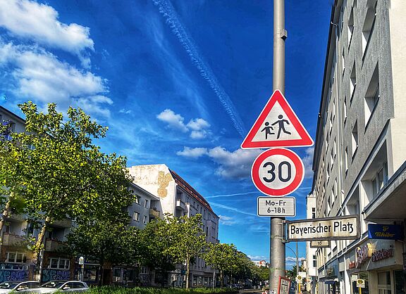 Auf dem Foto sind zwei Verkehrsschilder vor blauem Himmel zu sehen: Tempo 30 und Kinder. Rechts liegt eine Häuserzeile, auf der linken Seite ist die Straße durch Bäume begrünt.