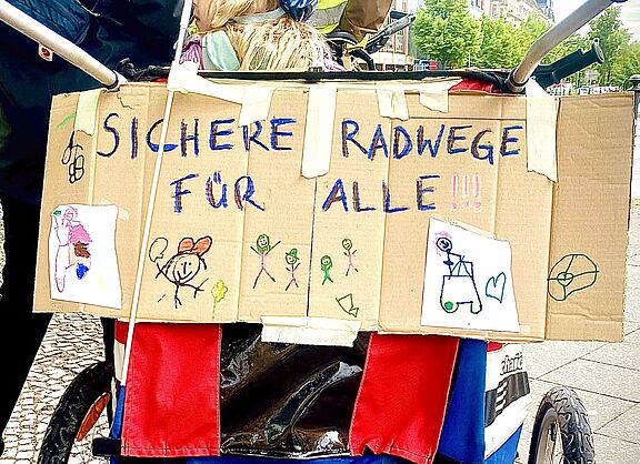 Auf dem Foto ist ein Pappschild zu seinen, dass an der Rückseite eines Fahrradanhängers angebracht ist. Neben bunten Kinderzeichnungen ist in gemalten Großbuchstaben „sichere Radwege für alle“ zu lesen. 