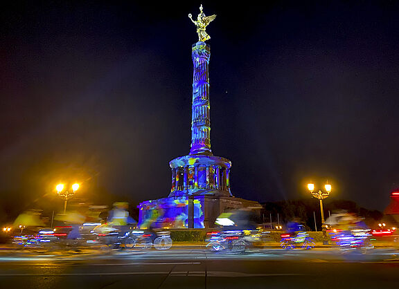 Menschen auf Fahrrädern fahren im Dunklen um die bunt angestrahlte Siegessäule