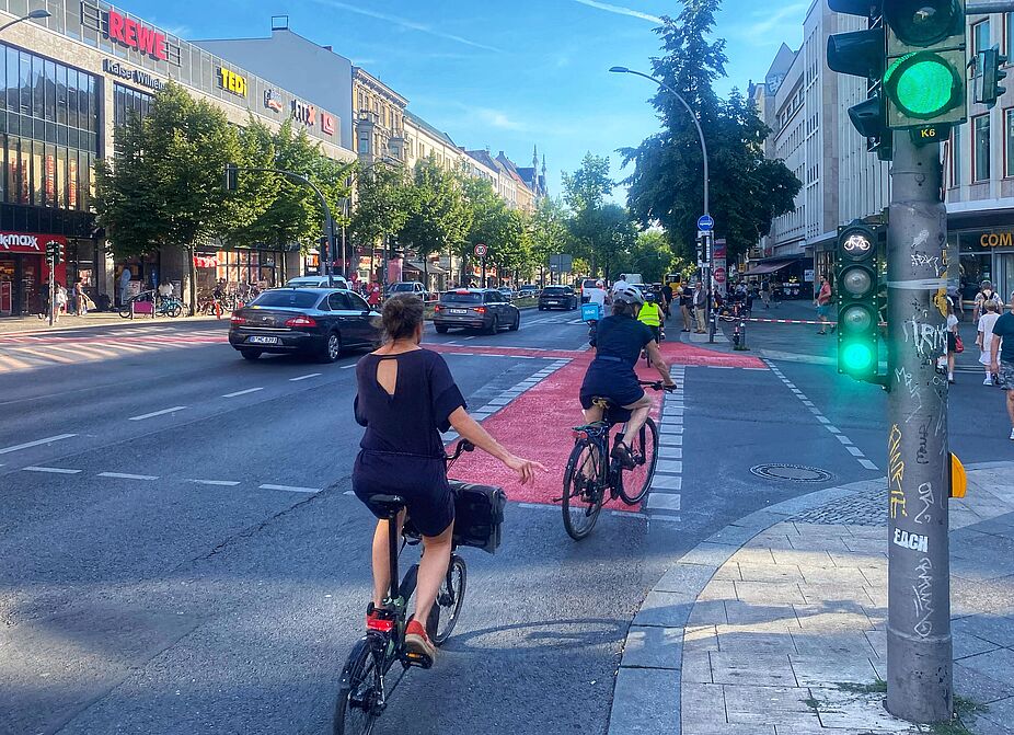 Juli 2024: Die Markierungsarbeiten am Richard-von-Weizsäcker-Platz Ecke Akazienstraße sind abgeschlossen. Der Radweg an der Kreuzung Akazienstraße ist rot markiert, und es fahren mehrere Menschen auf Fahrrädern dort entlang