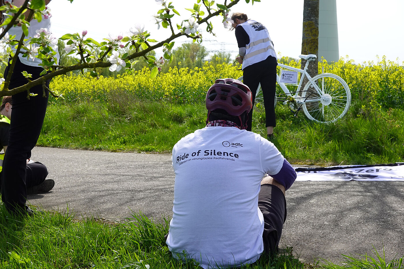 ein weißes Geisterrad wird am Umfallort am Wegrand aufgestellt. Im Vordergrund sitzt ein Mensch mit weißem Ride of Silence T-Shirt