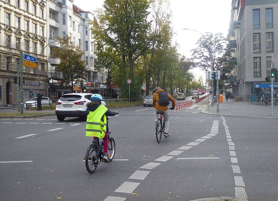 Die Hauptstraße hat zwischen Kleistpark und Dominicusstraße jetzt an beiden Seiten einen geschützten Radweg. Das Foto zeigt die Kreuzung am Kleistpark Hauptstraße Ecke Grunewaldstraße. Ein Fahrradfahrer fährt mit seinem Kind über die Kreuzung, auf die neue geschützte Radspur zu.