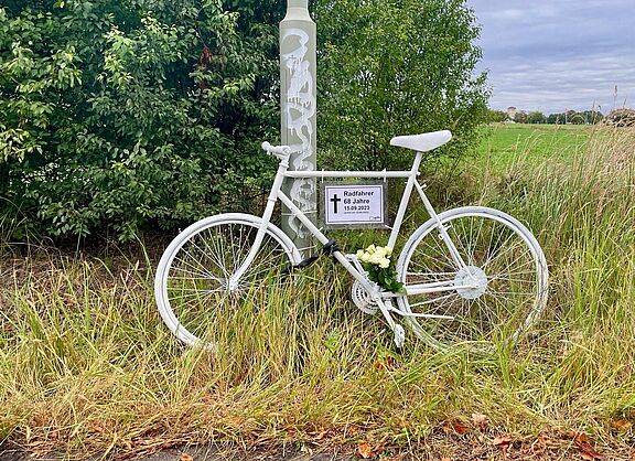 weißes Geisterrad mit Schild und weißen Rosen am Wegrand