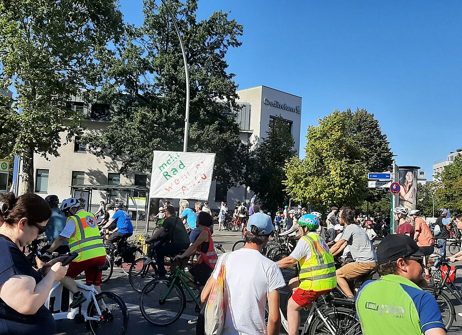 Kidical Mass Berlin und ADFC Kreisfahrt am Nollendorfplatz Am Nollendorfplatz steht eine Gruppe Menschen mit ihren Fahrrädern bei einer Demonstration. Der Himmel ist blau.