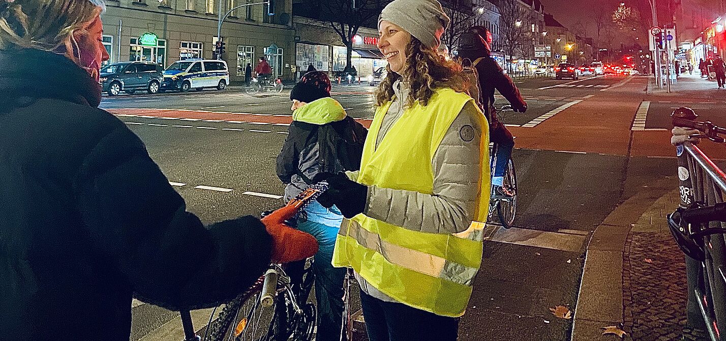 Eine Frau mit gelber Warnweste steht an einer roten Ampel in Berlin Schöneberg,  Hauptstraße Ecke Kolonnenstraße, und übergibt einer Radfahrerin eine Karte des ADFC mit einem Lebkuchen.