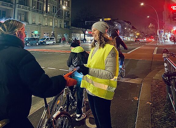 Eine Frau mit gelber Warnweste steht an einer roten Ampel in Berlin Schöneberg,  Hauptstraße Ecke Kolonnenstraße, und übergibt einer Radfahrerin eine Karte des ADFC mit einem Lebkuchen.