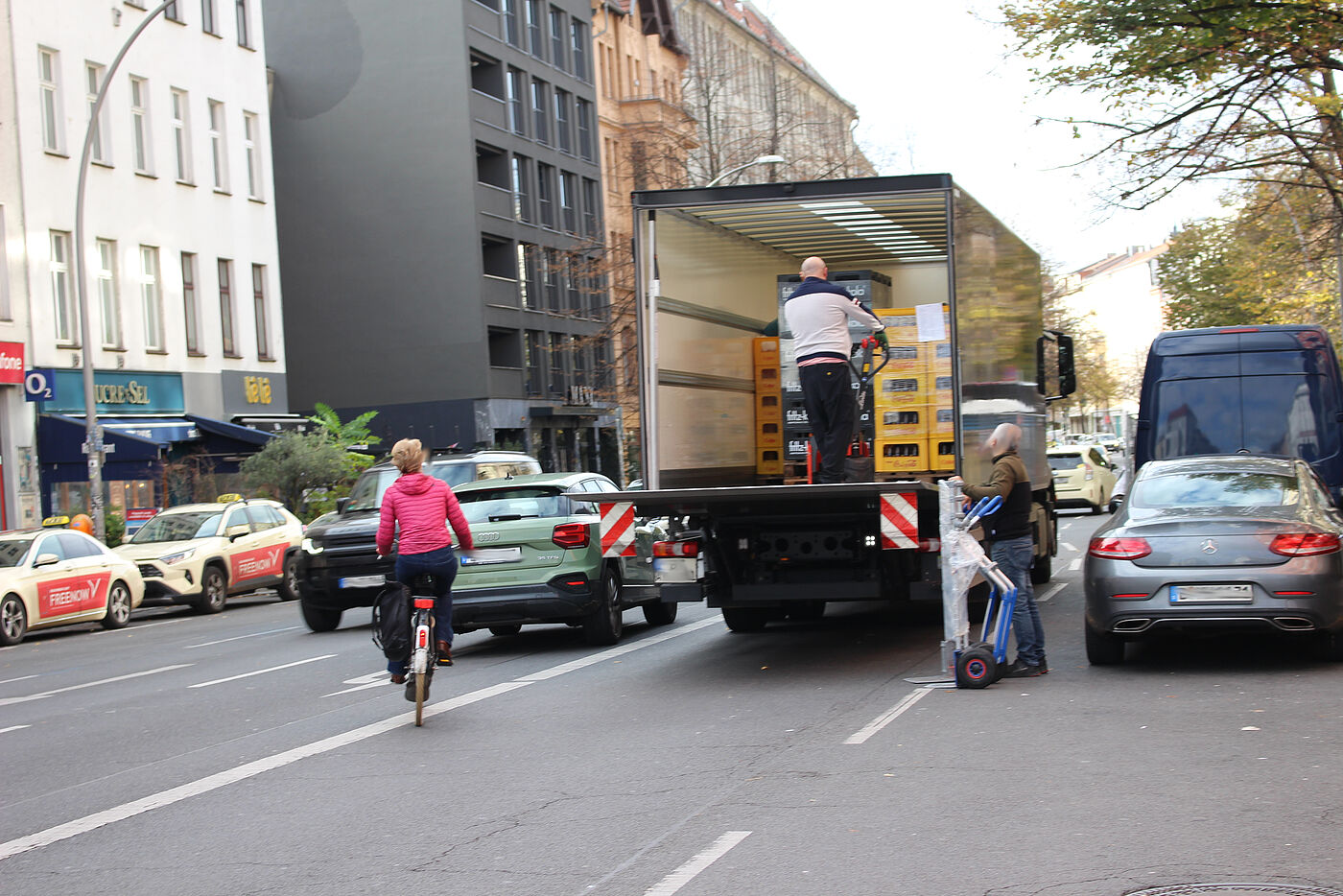 rechts Pkw und Lieferwagen, Mitte ein Lkw mit offener Ladeklappe, links davon Pkw und Radfahrerin weichen auf Gegenfahrbahn aus