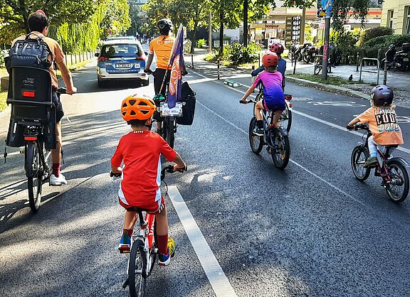 Eine Gruppe Kinder fährt bei sonnigem Wetter auf Fahrrädern das Berliner Reichpietschufer entlang, auf der Fahrbahn. Vorne fährt ein Auto der Polizei Berlin als Führungsfahrzeug, und zwei Erwachsene. Es ist eine ADFC Berlin Fahne zu sehen.