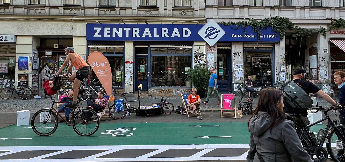 Ein Zweirad fährt über einen grünen Pop-Up Radweg, Menschen auf Liegestühlen im Hintergrund