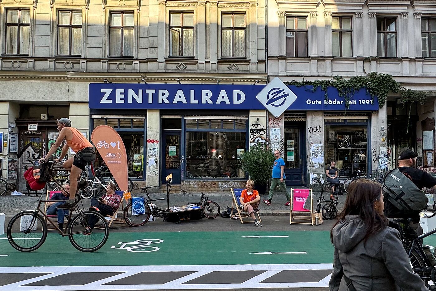 Ein Zweirad fährt über einen grünen Pop-Up Radweg, Menschen auf Liegestühlen im Hintergrund