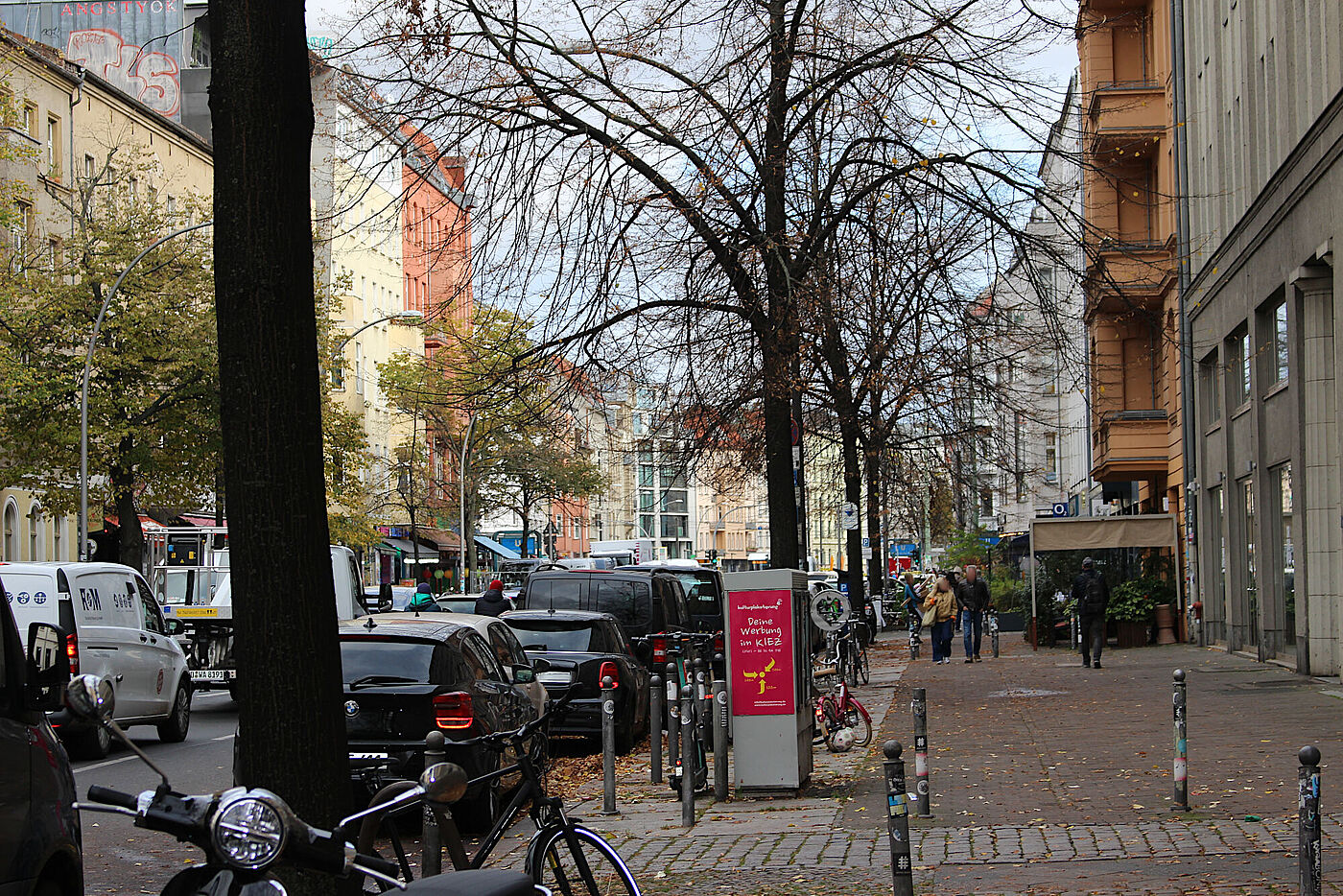Ansicht der Torstraße vom Gehweg aus, links Bäume, rechts Vorbauten von Gastronomie, dazwischen gerade mal Platz für 2 Fußgänger:innen