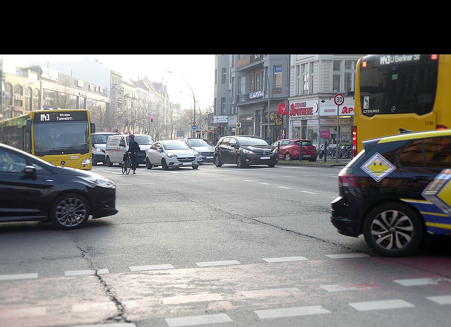Hauptstraße Schöneberg vor dem Umbau im März 2024 Das Foto zeigt die Hauptstraße Schöneberg am Richard-von-Weizsäcker-Platz im März 2024. Eine einzelne Radfahrerin steht an der Ampel und wirkt klein, angesichts vieler Autos und Busse, die um sie herum fahren. Auf der Fahrbahn ist kaum Platz.
