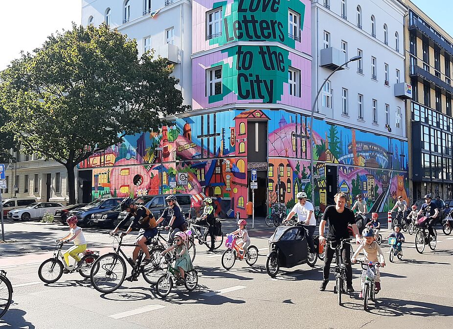 Kidical Mass Schöneberg 21.09.2024 auf der Bülowstraße vor dem Street Art Museum Eine Gruppe Erwachsener und Kinder fahren mit ihren Fahrrädern an einem bunt angemalten Haus auf der Bülowstraße vorbei.