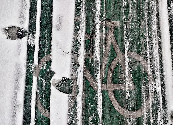 Man sieht eine grüne Bodenmarkierung eines Radwegs. Darüber liegt Schnee, der von Rad- und Fußabdrücken zerfurcht ist. 