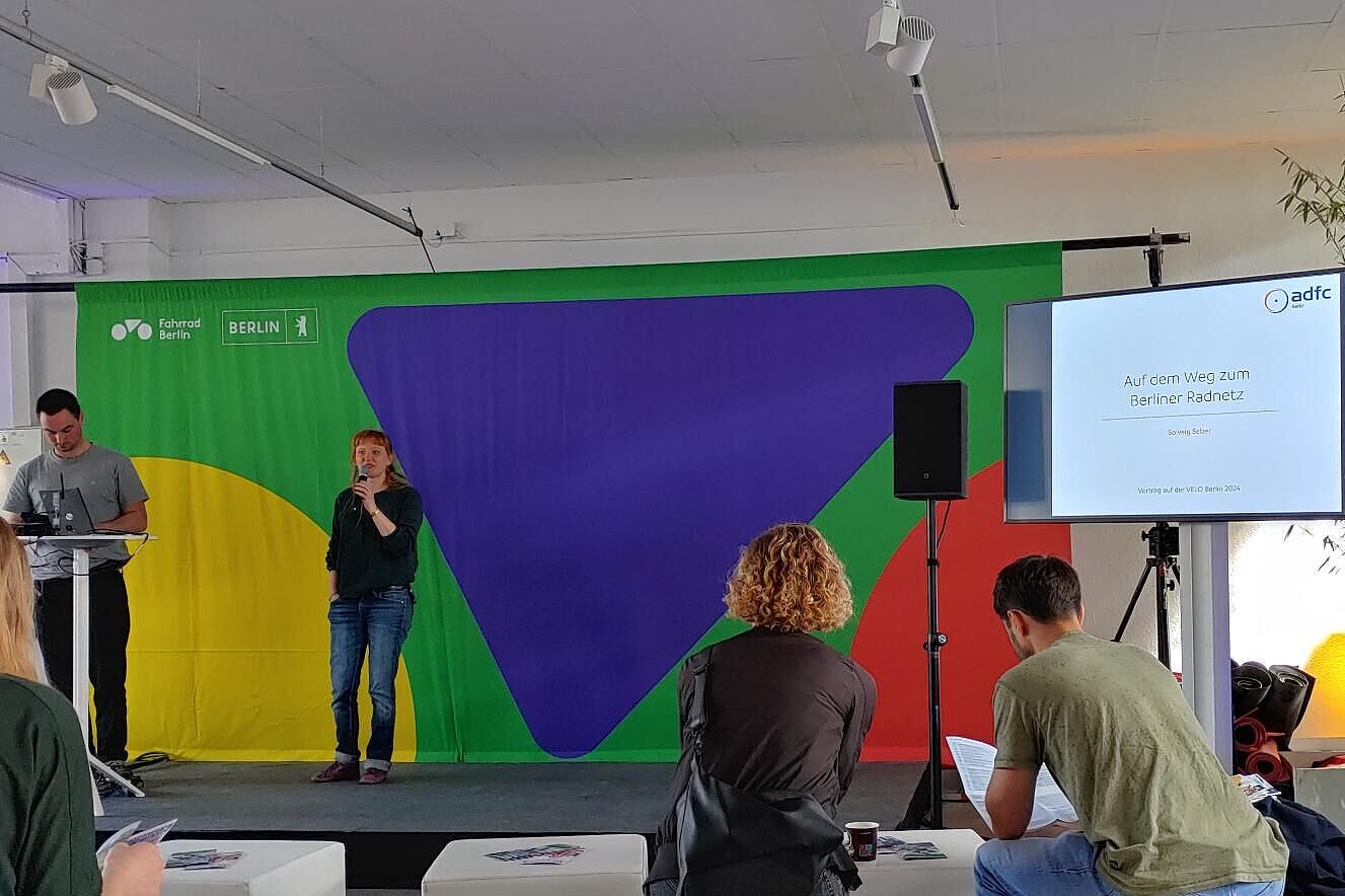 Menschen sitzen auf weißen Hockern und hören einer Referentin auf dem Podium zu