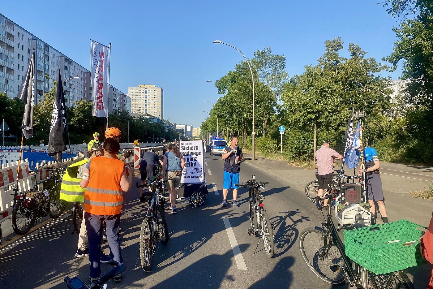 Eine Gruppe Radfahrender steht mit Fahnen auf der Landsberger Allee und hört einem Sprecher zu, der eine Zwischenkundgebung hält.