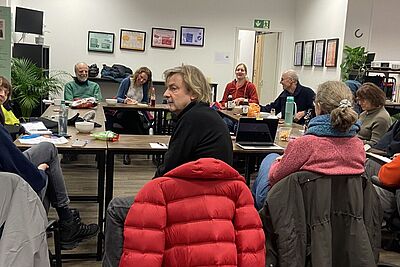 Eine Gruppe Menschen sitzt in einem großen, hell erleuchteten Raum um Tische, die im Kreis aufgestellt sind.