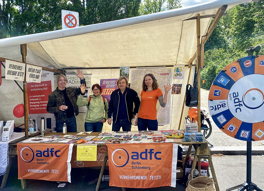 Stand des ADFC Schöneberg und ADFC Tempelhof beim Nachbarschaftsfest am Schöneberger Rathaus am 24.05.2025 mit Glücksrad. Stand des ADFC Schöneberg und Tempelhof beim Nachbarschaftsfest am Schöneberger Rathaus am 24.05.2025 mit Glücksrad und vier Standbetreuer:innen, die winken. Es sind Fahnen, Plakate und Beachflags des ADFC Berlin in Orangetönen zu sehen.