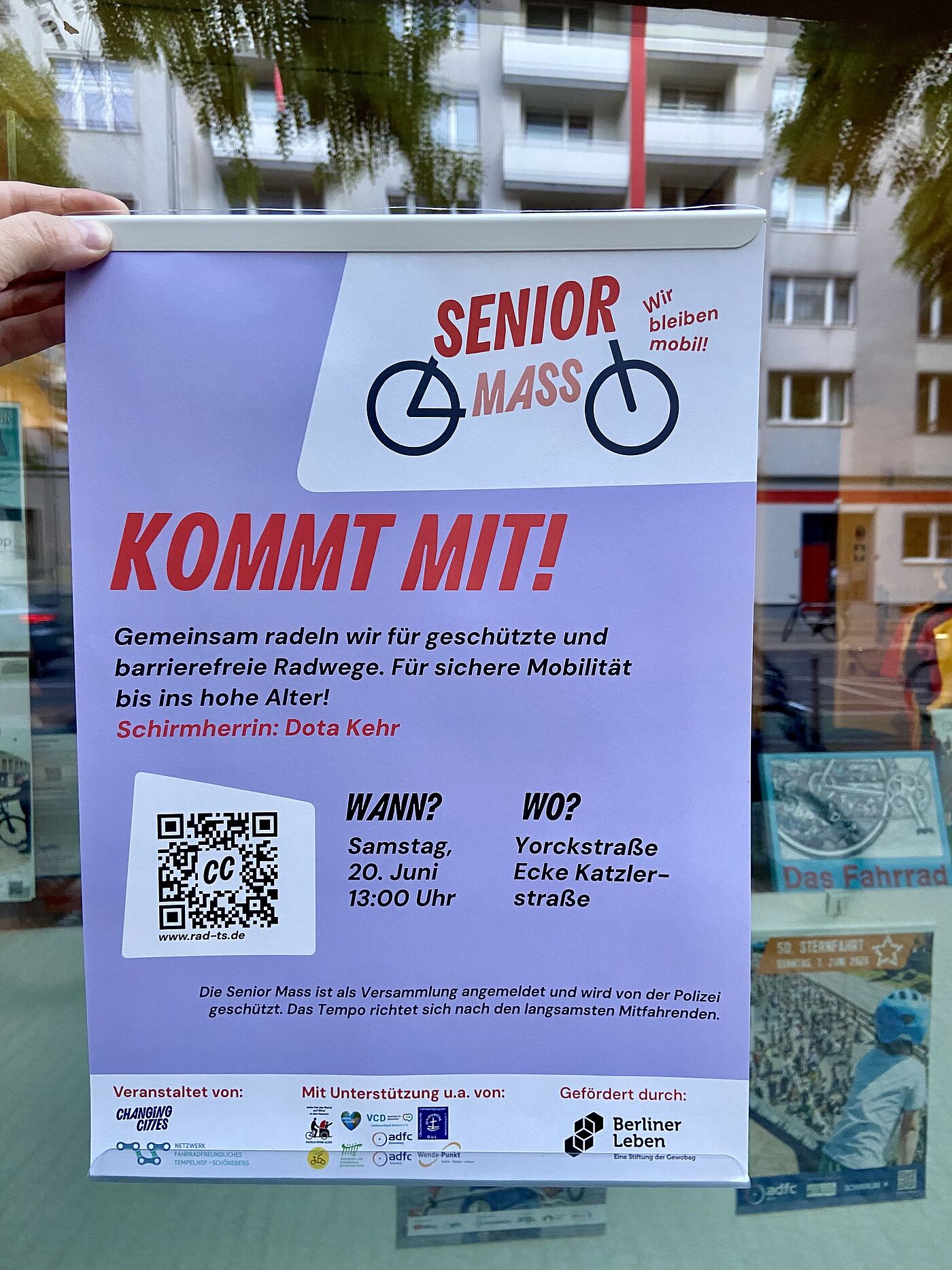 Plakat von Changing Cities wird vor einer Schaufensterscheibt des ADFC Berlin VeloKiez hochgehalten