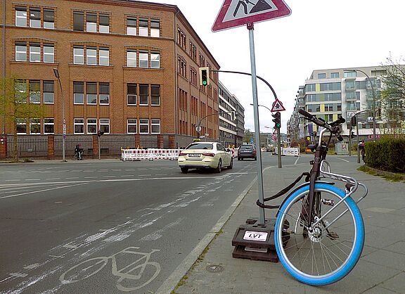 Auf der Ella-Barowsky-Straße in Schöneberg fahren Richtung Bahnhof Südkreuz Autos. Es ist eine Kreuzung zu sehen, ein schmaler Radweg mit Piktogram, sowie ein Fahrrad, das am Ständer eines Baustellenschildes angeschlossen ist. 