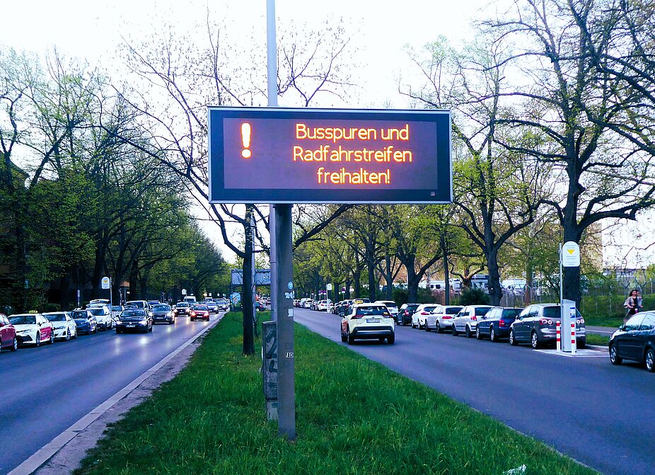 Mehr Verkehrssicherheit am Tempelhofer Damm? Gern SenMVKU, und lasst dabei doch bitte die Bäume stehen! Foto vom Tempelhofer Damm, vom begrünten Mittelstreifen aus Richtung Süden betrachtet. Rechts und links fahren auf jeweils zwei Fahrspuren Autos. Sowohl auf dem Mitteistreifen als auch am Fahrbahnrand stehen Bäume. Ein großes Schild mit beleuchteter Schrift fordert Autofahrende dazu auf, Maßnahmen für mehr Verkehrssicherheit einzuhalten: Rad- und Busstreifen freihalten.