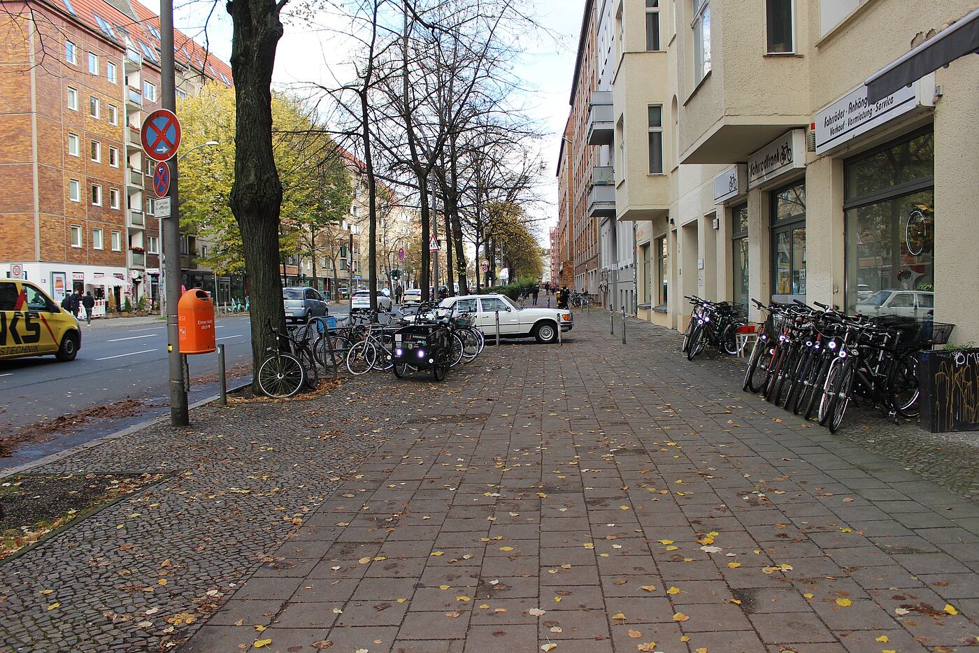 breiter Gehweg einer Stadtstraße, rechts querstehende Fahrräder vor einem Fahrradgeschäft, links geparkte Fahrräder, ein querstehendes Auto in einer Einfahrt, im Hintergrund Fußgänger:innen