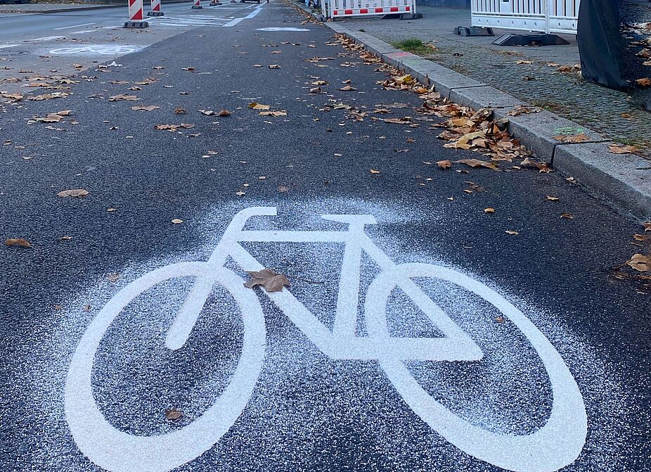November/Dezember 2024: Ein Fahrrad-Piktogramm kennzeichnet den neuen geschützten Radstreifen ab Martin-Luther-Straße Richtung Bayerischer Platz. Auf einer frisch asphaltierten Fläche ist auf der rechten Fahrbahn der Grunewaldstraße ein Fahrradpiktogramm zu sehen. Es ist frisch aufgemalt, was am strahlenden Weiß und den Steinchen zum Trocknen zu sehen ist. Das Piktogramm befindet sich zwischen Martin-Luther-Straße und Berchtesgadenerstraße Richtung Bayerischer Platz