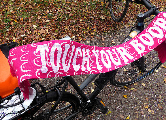 An einem Fahrrad hängt ein rosa Schal mit Brustsymbolen und der Schrift "Touch your Boobs" als Analogie der Selbstvorsorge.