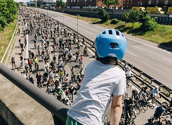 Kind mit blauem Helm guckt auf Autobahn über die auf einer Seite hunderte FahrradfahrerInnen fahren