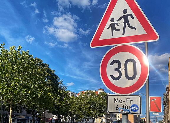 Auf dem Foto sind zwei Verkehrsschilder vor blauem Himmel zu sehen: Tempo 30 und Kinder. Rechts liegt eine Häuserzeile, auf der linken Seite ist die Straße durch Bäume begrünt.