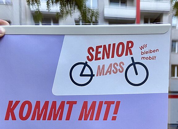 Ein Plakat mit dem Titel „Senior Mass – Kommt mit!“ wird vor einer Schaufensterscheibe ins Licht gehalten.