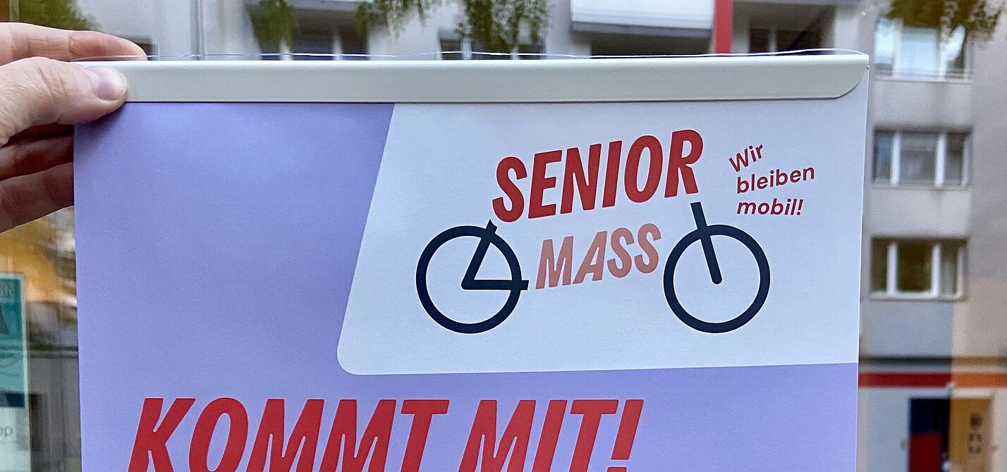 Ein Plakat mit dem Titel „Senior Mass – Kommt mit!“ wird vor einer Schaufensterscheibe ins Licht gehalten.