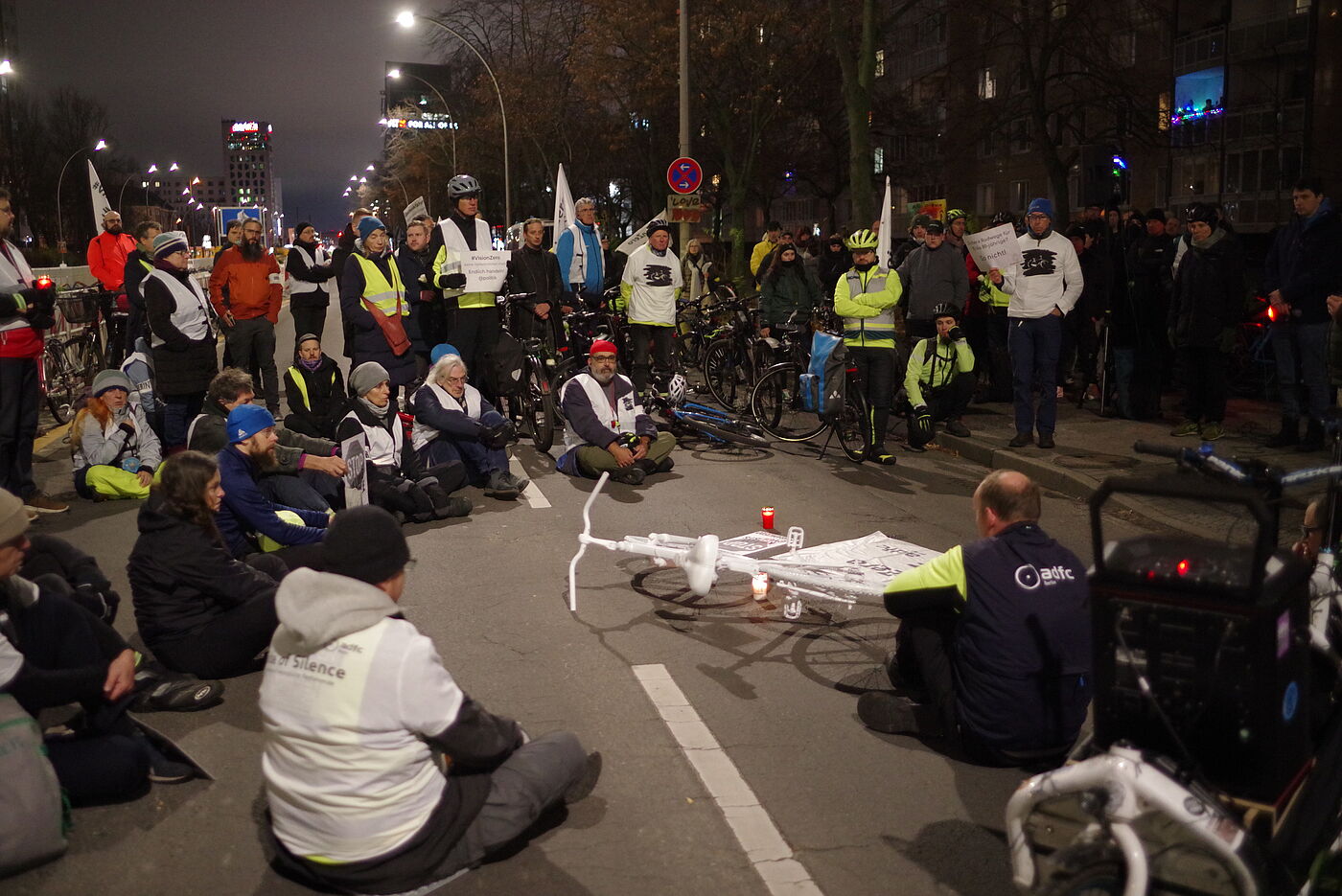 Menschen sitzen bei einer Mahnwache um ein weißes Geisterrad auf der Straße