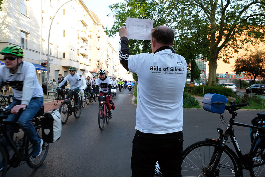 Ein Mann hält ein Schild hoch, im Hintergrund fahren Radfahrende vorbei, Ride of Silence 2023 in Berlin.