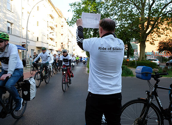 Ein Mann hält ein Schild hoch, im Hintergrund fahren Radfahrende vorbei, Ride of Silence 2023 in Berlin.