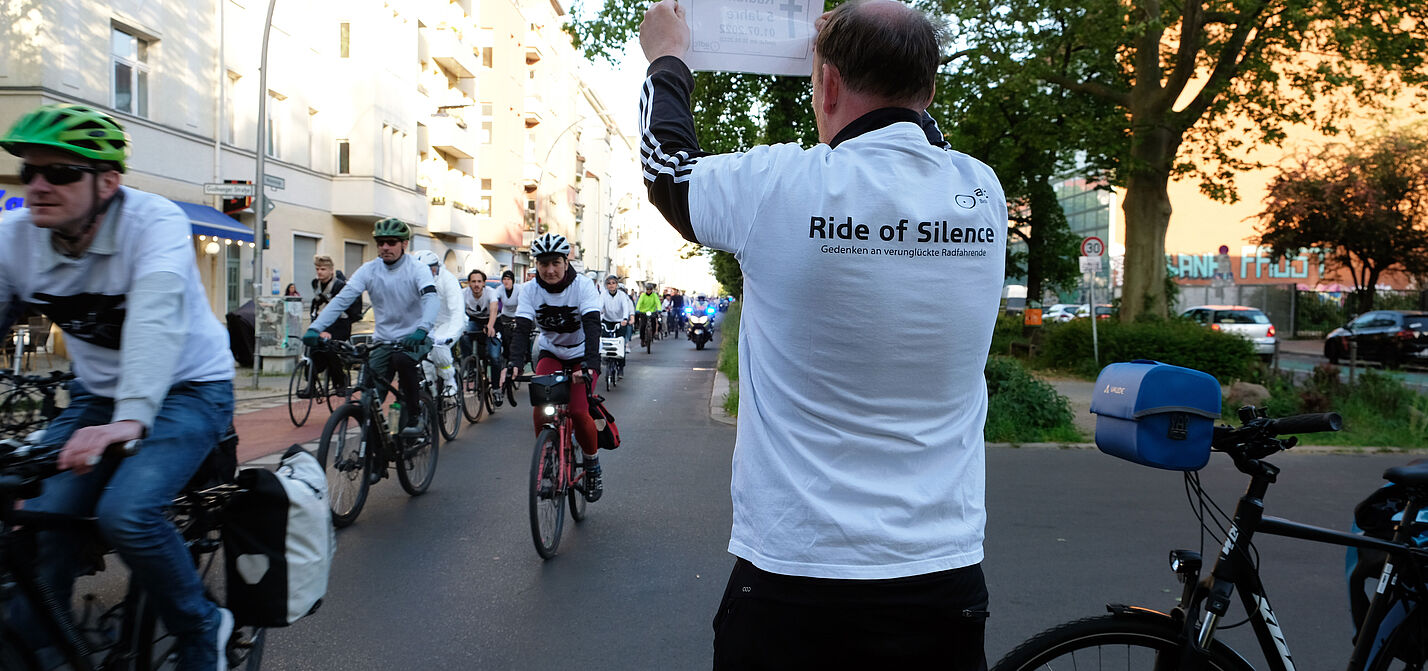 Ein Mann hält ein Schild hoch, im Hintergrund fahren Radfahrende vorbei, Ride of Silence 2023 in Berlin.