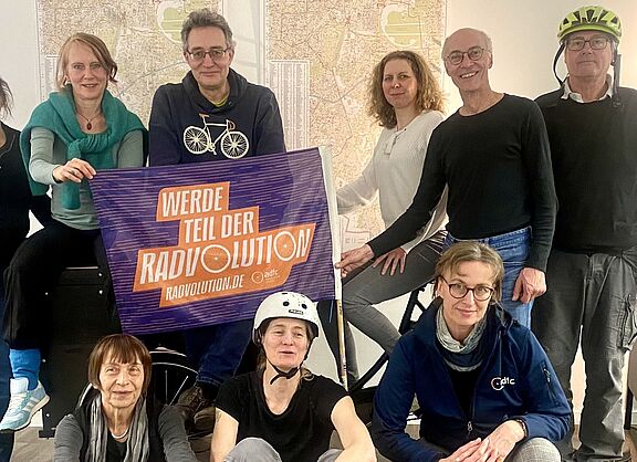 Eine Gruppe Menschen stehen um und sitzen vor und auf einem Lastenrad, einige tragen Fahrradhelme. Eine ADFC Fahne mit Schrift "Werde Teil der Radvolution" ist im Mittelpunkt. Im Hintergrund sind Karten von Berlin Schöneberg sichtbar, aber nicht zu entziffern.