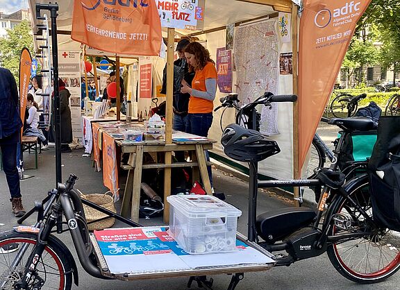 Stand des ADFC Schöneberg und Tempelhof beim Nachbarschaftsfest am Schöneberger Rathaus am 24.05.2025. Im Vordergund ist ein e-Lastenrad zu sehen. Auf der Ladefläche befindet sich Material für den Stand. Es sind Fahnen, Plakate und Beachflags des ADFC Berlin in Orangetönen zu sehen, und Personen, die den Stand betreuen. 