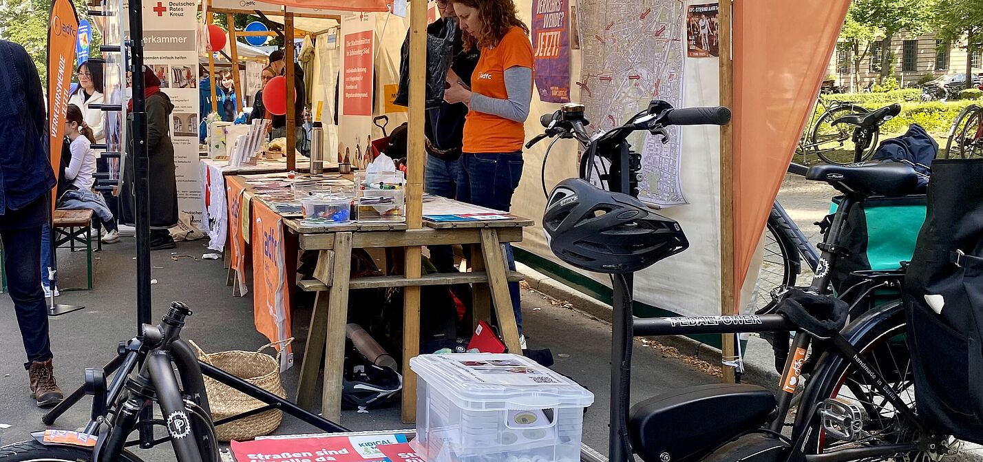 Stand des ADFC Schöneberg und Tempelhof beim Nachbarschaftsfest am Schöneberger Rathaus am 24.05.2025 mit Lastenrad e-Harry Stand des ADFC Schöneberg und Tempelhof beim Nachbarschaftsfest am Schöneberger Rathaus am 24.05.2025. Im Vordergund ist ein e-Lastenrad zu sehen. Auf der Ladefläche befindet sich Material für den Stand. Es sind Fahnen, Plakate und Beachflags des ADFC Berlin in Orangetönen zu sehen, und Personen, die den Stand betreuen.