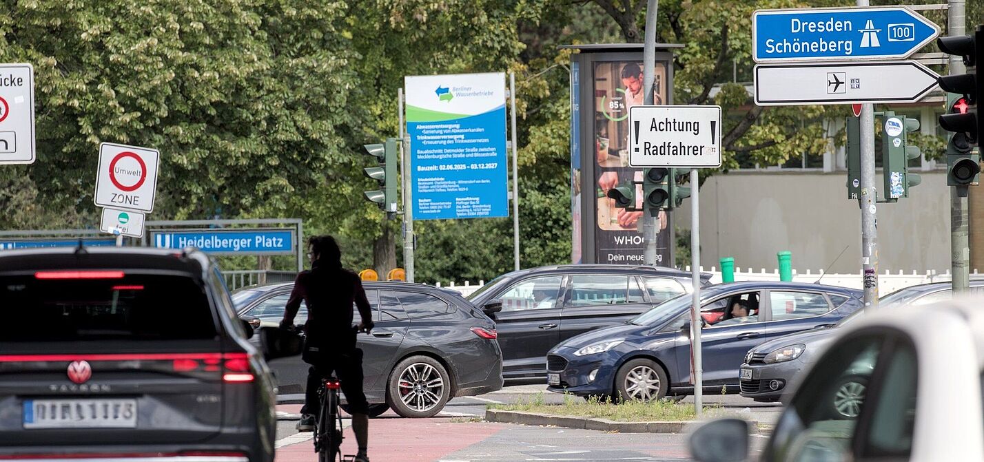 Auf einer Hauptverkehrsstraße am Heidelberger Platz in Berlin ist unter einer Brücke nahe einer Auffahrt zur A100 die Silhoutte eines Radfahrers zu sehen. Er steht mit seinem Rad mitten auf der Fahrbahn, umgeben von Autos. Zum Radfahren ist dort wenig Platz. Es sind mehrere Schilder zu sehen, unter anderem ein weißes Schild, das die Aufschrift „Achtung Radfahrer“ trägt.
