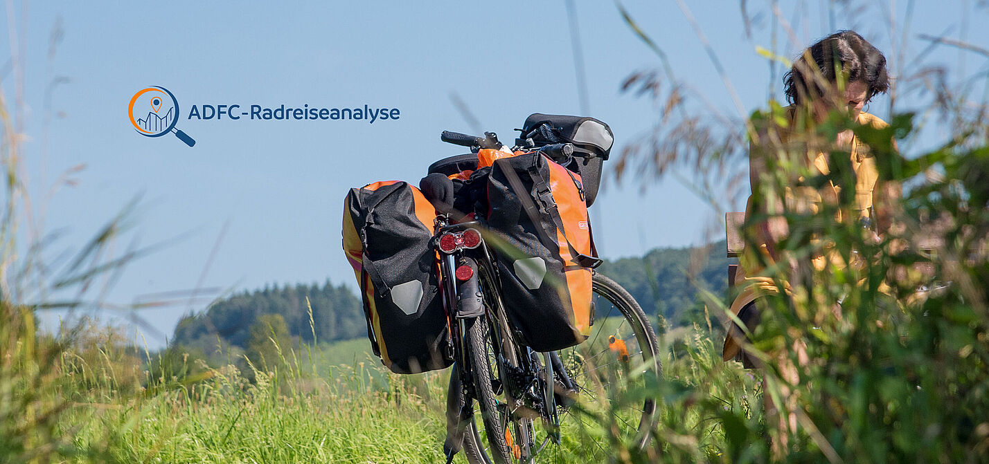 ADFC-Radreiseanalyse 2023 Reiserad mit Packtaschen steht in einer gemähten Wiese, Frau hinter hochgewachsenem Gras